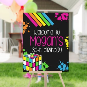 May include: A black sign on an easel with the text "Welcome to Megan's 30th birthday" in white and pink. The sign has colorful paint splatters and a Rubik's Cube graphic. Balloons are visible in the background.