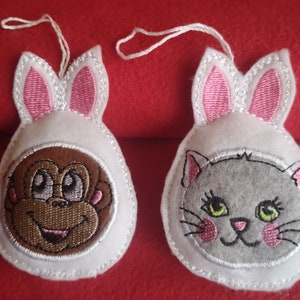 May include: Two felt ornaments, one featuring a monkey and the other a cat, both wearing bunny ear hoods. The monkey has brown fur and a smiling face. The cat is gray with pink cheeks and green eyes. Each ornament has a white string for hanging.