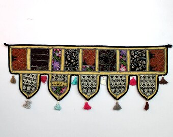 May include: A colorful patchwork fabric door hanging with tassels. The fabric is in shades of black, brown, gold, purple, and white. The door hanging has a scalloped bottom edge and is decorated with embroidered details.