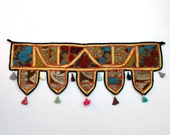 May include: A colorful fabric door hanging with a patchwork design and tassels. The hanging is made of various fabrics in shades of brown, yellow, red, blue, and green. The tassels are in shades of pink, blue, and green.