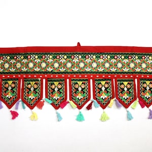 Może przedstawiać: Wiszący ozdóbny element do drzwi w kolorze czerwonym, zielonym i złotym, z kolorowymi frędzlami. Ozdoba ma geometryczny wzór i zdobienia przypominające lustra.