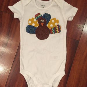 Puede incluir: Body de beb&eacute; blanco con un dise&ntilde;o de pavo colorido. El pavo tiene un cuerpo marr&oacute;n, un pico rojo y plumas estampadas de colores en amarillo, azul y rojo. El body es talla 18 meses.