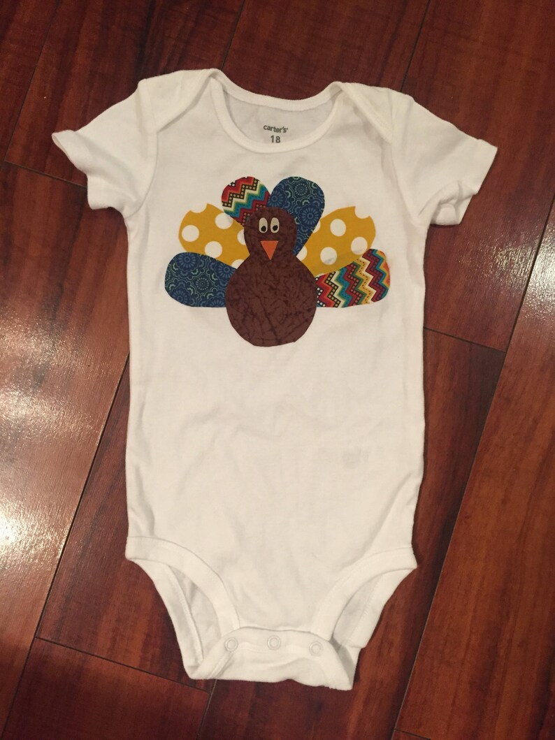 Puede incluir: Body de beb&eacute; blanco con un dise&ntilde;o de pavo colorido. El pavo tiene un cuerpo marr&oacute;n, un pico rojo y plumas estampadas de colores en amarillo, azul y rojo. El body es talla 18 meses.