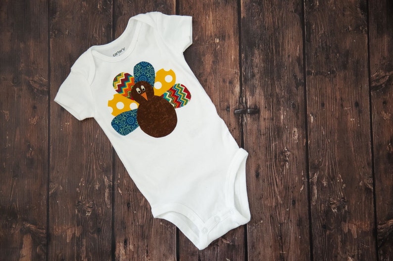 Puede incluir: Body de beb&eacute; blanco con un dise&ntilde;o de pavo colorido. El pavo tiene un cuerpo marr&oacute;n, un pico amarillo y una cola vibrante con patrones azules, amarillos y rojos. El body est&aacute; hecho de material suave.