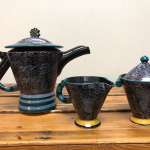 Könnte beinhalten: Ein schwarzes und türkisfarbenes Keramik-Set aus Teekanne, Milchkännchen und Zuckerdose mit einem gesprenkelten Überzug. Die Teekanne hat einen einzigartigen Griff und einen Deckel mit einem dekorativen Knopf. Das Milchkännchen und die Zuckerdose haben passende Griffe und Deckel.