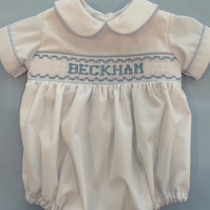 May include: A white baby onesie with blue trim and a blue embroidered name 'BECKHAM'.