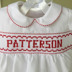 May include: White baby dress with a Peter Pan collar and red trim. The dress features the name "PATTERSON" embroidered in red across the chest. A white satin ribbon is tied in a bow at the neckline.