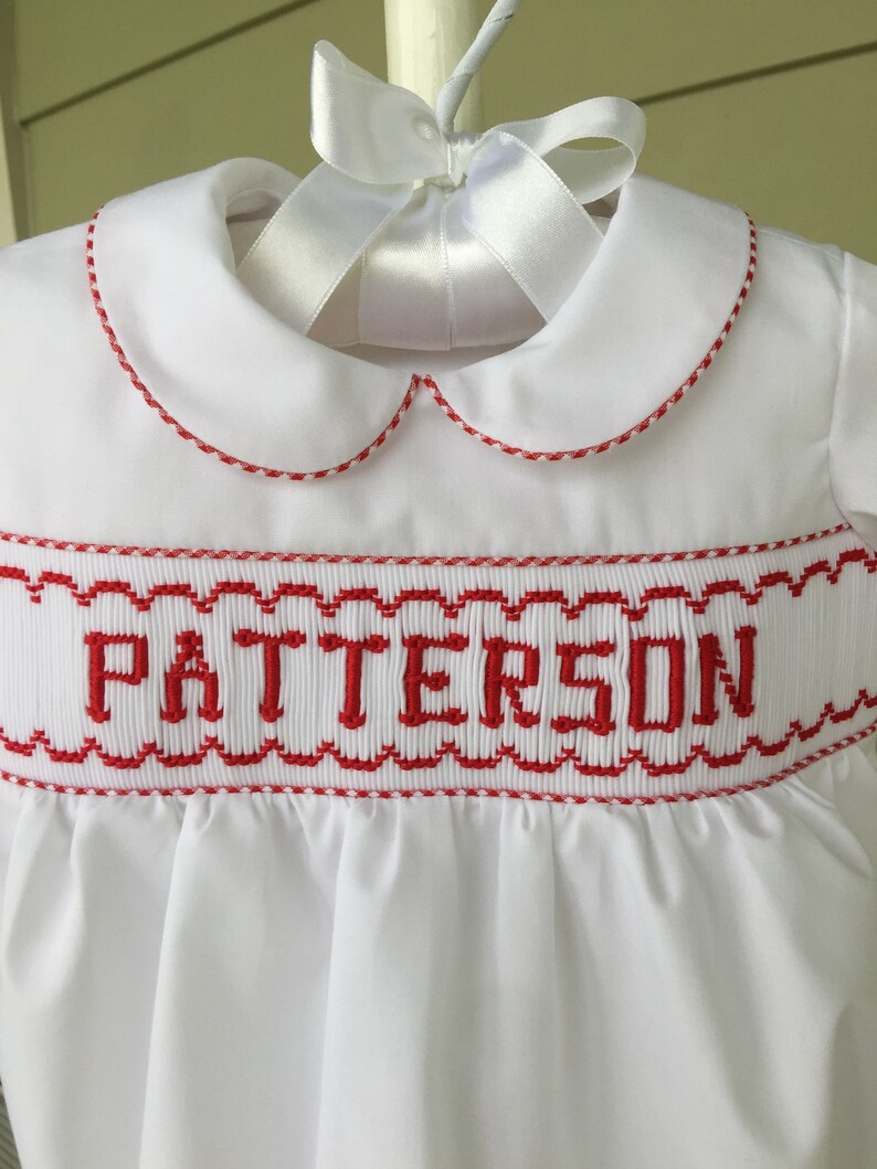 May include: White baby dress with a Peter Pan collar and red trim. The dress features the name "PATTERSON" embroidered in red across the chest. A white satin ribbon is tied in a bow at the neckline.