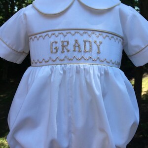 May include: White baby romper with a Peter Pan collar and short sleeves. The romper features gold embroidery with the name "GRADY" across the chest. The edges of the collar and sleeves are trimmed with gold. The garment has a gathered, bubble-style bottom.