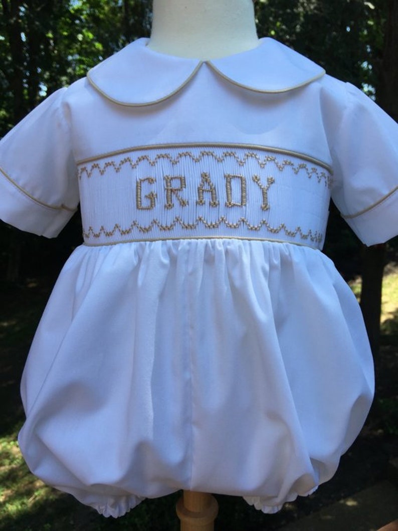 May include: White baby romper with a Peter Pan collar and short sleeves. The romper features gold embroidery with the name "GRADY" across the chest. The edges of the collar and sleeves are trimmed with gold. The garment has a gathered, bubble-style bottom.
