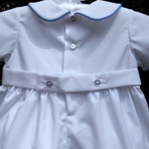 May include: White baby romper with a blue trim on the collar and sleeves. The romper has a button-down front and a matching belt with buttons. The outfit is hanging on a white hanger with a white bow.