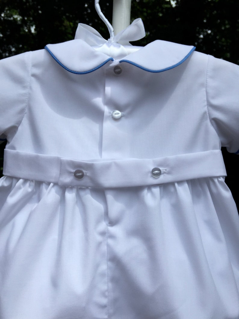May include: White baby romper with a blue trim on the collar and sleeves. The romper has a button-down front and a matching belt with buttons. The outfit is hanging on a white hanger with a white bow.