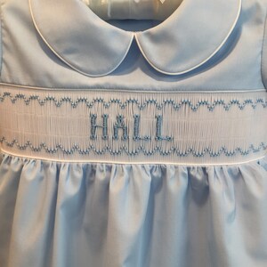 May include: A light blue baby dress with a white collar and trim. The dress features smocking with the name "HALL" embroidered in blue thread. The dress is hanging on a white hanger with a white bow.