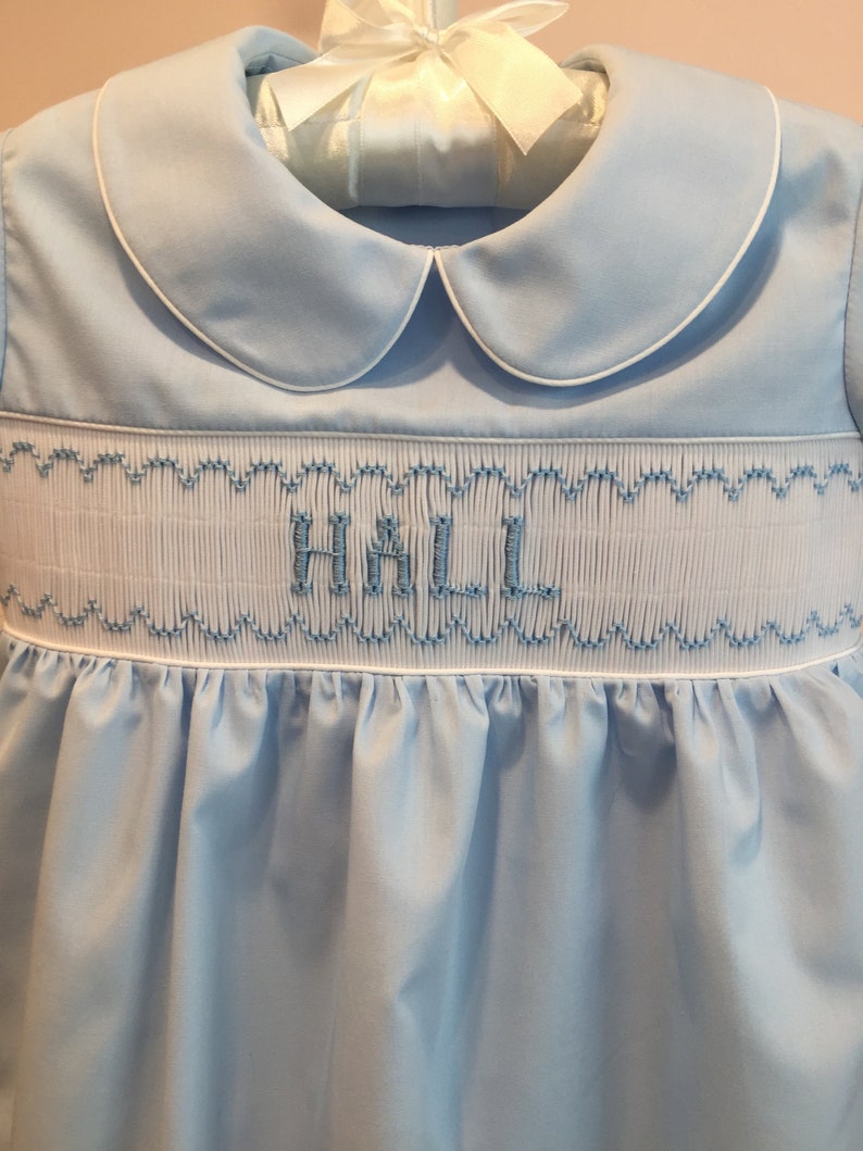 May include: A light blue baby dress with a white collar and trim. The dress features smocking with the name "HALL" embroidered in blue thread. The dress is hanging on a white hanger with a white bow.