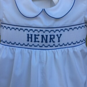 May include: White baby romper with blue trim and the name "HENRY" embroidered across the chest. The romper features a Peter Pan collar and a ribbon tie at the back. The design includes a smocked waistline with blue stitching.