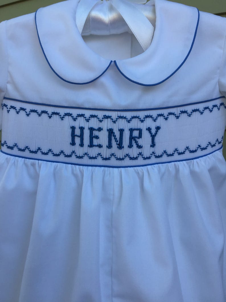 May include: White baby romper with blue trim and the name "HENRY" embroidered across the chest. The romper features a Peter Pan collar and a ribbon tie at the back. The design includes a smocked waistline with blue stitching.