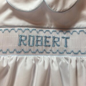 May include: White baby romper with a Peter Pan collar and light blue trim. The word "ROBERT" is embroidered in light blue thread across the chest. The romper features smocking and a gathered skirt.