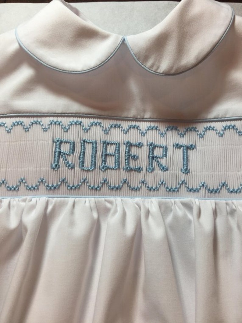 May include: White baby romper with a Peter Pan collar and light blue trim. The word "ROBERT" is embroidered in light blue thread across the chest. The romper features smocking and a gathered skirt.