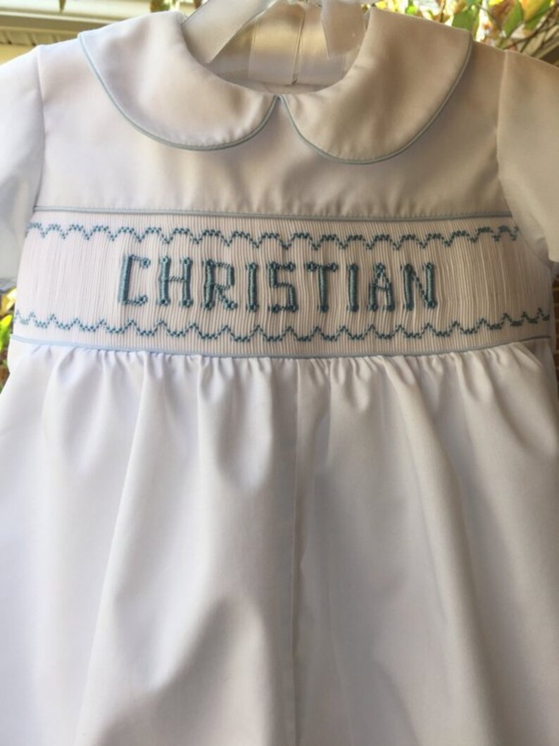 May include: White baby romper with a Peter Pan collar and light blue trim. The word "CHRISTIAN" is embroidered in light blue across the chest. The romper is hanging on a white hanger.