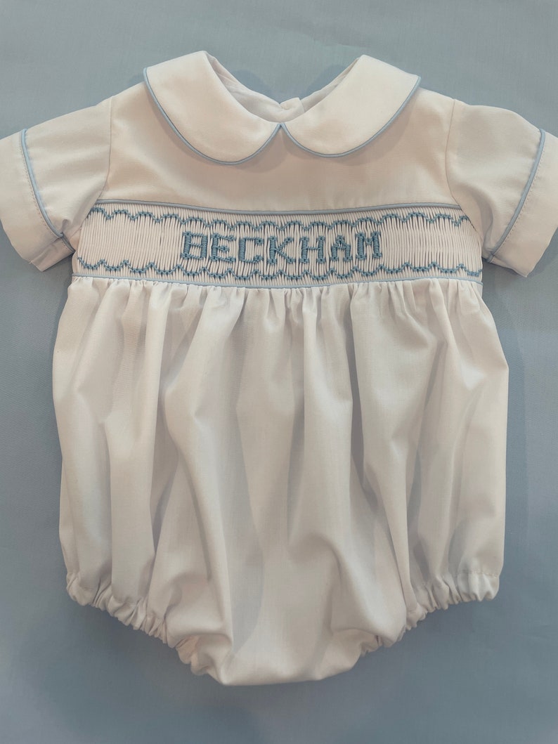 May include: A white baby onesie with blue trim and a blue embroidered name 'BECKHAM'.