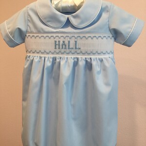 May include: Light blue baby bubble with white trim and a Peter Pan collar. The bubble features smocking across the chest with the name "HALL" embroidered in blue thread. The garment is hanging on a white hanger.