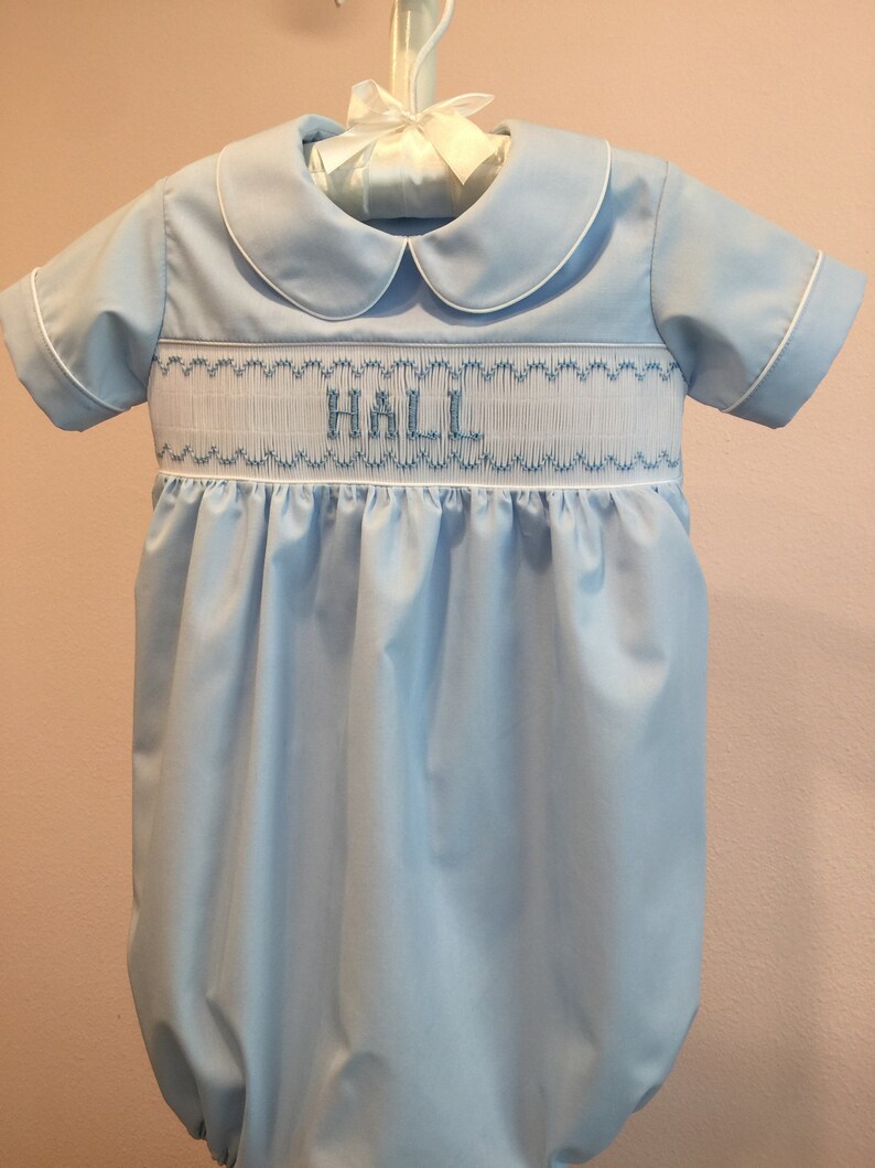 May include: Light blue baby bubble with white trim and a Peter Pan collar. The bubble features smocking across the chest with the name "HALL" embroidered in blue thread. The garment is hanging on a white hanger.