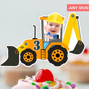 Może przedstawiać: Żółty plastikowy pojazd budowlany z czarną łyżką, numerem 3 z boku i twarzą dziecka na siedzeniu kierowcy. Pojazd jest na patyku, co czyni go ozdobą tortu.