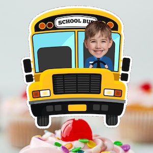 May include: A yellow school bus with a smiling child's face in the driver's seat. The bus has the words "SCHOOL BUS" on the front.