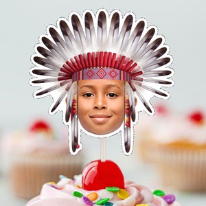 May include: A red, white, and black paper cutout of a child wearing a headdress with feathers. The cutout is on a stick and is placed on top of a cupcake.