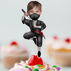 Puede incluir: Una ilustración de dibujos animados de un niño vestido de ninja, con una máscara negra y sosteniendo una espada. La ilustración está en un adorno para pastel.