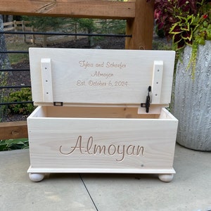 May include: A white wooden toy chest with a hinged lid. The lid is open and has the text "Tykeia and Schaefer Almoyan Est. October 6, 2024" engraved on it. The front of the chest has the name "Almoyan" engraved on it.
