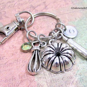 May include: Silver-toned keychain with a lobster clasp, key ring, and multiple charms. Charms include a whisk, a bundt cake, a rolling pin, a small round charm, and a green gemstone. The keychain is designed with baking-themed charms.