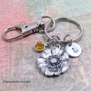 May include: A silver-toned keychain with a flower charm, a round initial charm, and a yellow gemstone. The flower charm has detailed petals and a central gem. The keychain includes a clasp and a split ring.