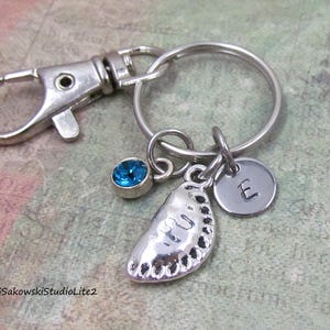 May include: A silver-toned metal keychain featuring a lobster clasp and a circular ring. Attached to the ring are three charms: a small blue crystal, a silver-toned dumpling-shaped charm, and a round silver-toned disc stamped with the letter E.