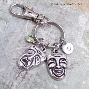 May include: Silver-toned keychain with a lobster clasp, a split ring, and charms. Charms include a green gemstone, a smiling theater mask, a frowning theater mask, and a small disc with the letter "D".