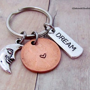 May include: A silver key chain with a silver crescent moon charm, a copper penny with a heart engraved on it, and a silver tag that says "DREAM".