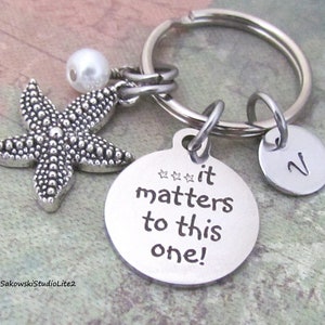 May include: Silver keychain with a starfish charm, a pearl bead, and a round tag that says "it matters to this one!" with four stars above the text. There is also a small round tag with the letter "N" on it.