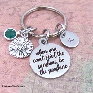May include: Silver keychain with a round charm that says "when you can't find the sunshine, be the sunshine". The keychain also has a small sunflower charm and a green gemstone.