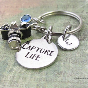 May include: A silver keychain with a small camera charm, a round charm that says "CAPTURE LIFE", and a smaller round charm with the letter "E".