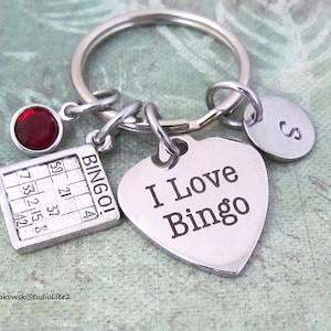 May include: A silver heart-shaped keychain with the text "I Love Bingo" engraved on it. The keychain also features a small silver bingo card charm, a red gemstone charm, and a silver charm with the letter "S" engraved on it.