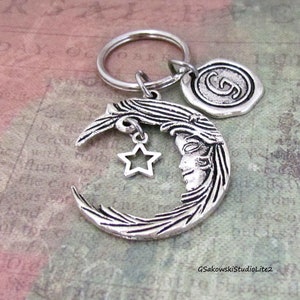 May include: Silver-toned crescent moon keychain with a face, a dangling star charm, and a circular charm with a letter. The keychain is attached to a silver ring. The background is a textured, multi-colored surface.