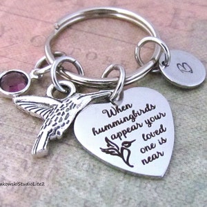 May include: A silver keychain with a hummingbird charm, a heart-shaped pendant with the text "When hummingbirds appear your loved one is near", a small round pendant with a letter "B", and a purple gemstone. The keychain is on a silver ring.