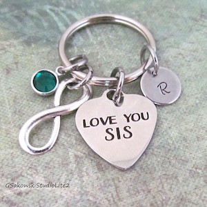 May include: Silver keychain with a heart-shaped charm that reads "LOVE YOU SIS." The keychain also features an infinity symbol, a small round charm with the letter "R," and a green gemstone.