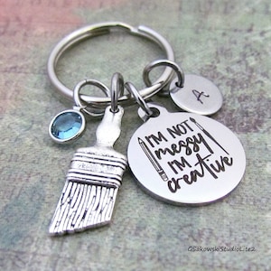 May include: Silver keychain with a paintbrush charm, a blue crystal charm, and a round tag that says "I'm not messy I'm creative" with a pencil and paintbrush graphic. The keychain also has a small round tag with the letter "A" on it.
