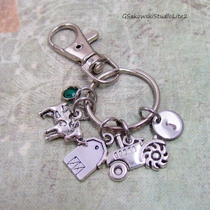 May include: Silver-toned keychain with a lobster clasp, a keyring, and several charms. Charms include a cow, a tractor, a tag, a green gem, and a small round charm. The keychain is a farm-themed accessory.