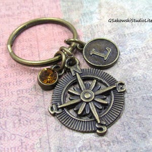 May include: Antique brass-colored keychain featuring a compass rose charm, a round charm with the letter "L", and a small amber-colored gemstone. The keychain is designed with a vintage aesthetic.