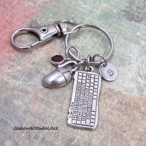 May include: Silver-toned keychain with a lobster clasp, a keyboard charm, a computer mouse charm, a small heart charm, and a purple gemstone. The keyboard charm has letters and numbers. The keychain is on a patterned background.