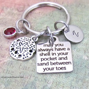 May include: Silver keychain with a charm that reads "may you always have a shell in your pocket and sand between your toes". The keychain also features a small red gemstone, a sand dollar charm, and a charm with the letter "N".