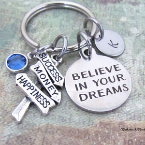 May include: Silver keychain with a blue crystal charm and three silver charms with the words "Happiness", "Money", and "Success" on them. The largest charm is a silver circle with the words "Believe in your dreams" on it.