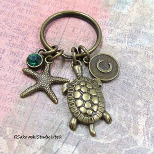 May include: Bronze-toned keychain featuring a starfish charm with a green gem, a turtle charm, and a circular charm with a letter "C". The keychain is designed with a nautical theme.